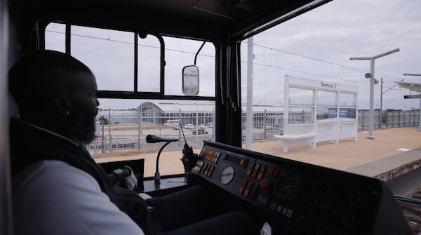 Interior shot of a MetroLink operator driving a train by the Lambert Airport Terminal #2 station. The platform of the station can be seen through the windshield.